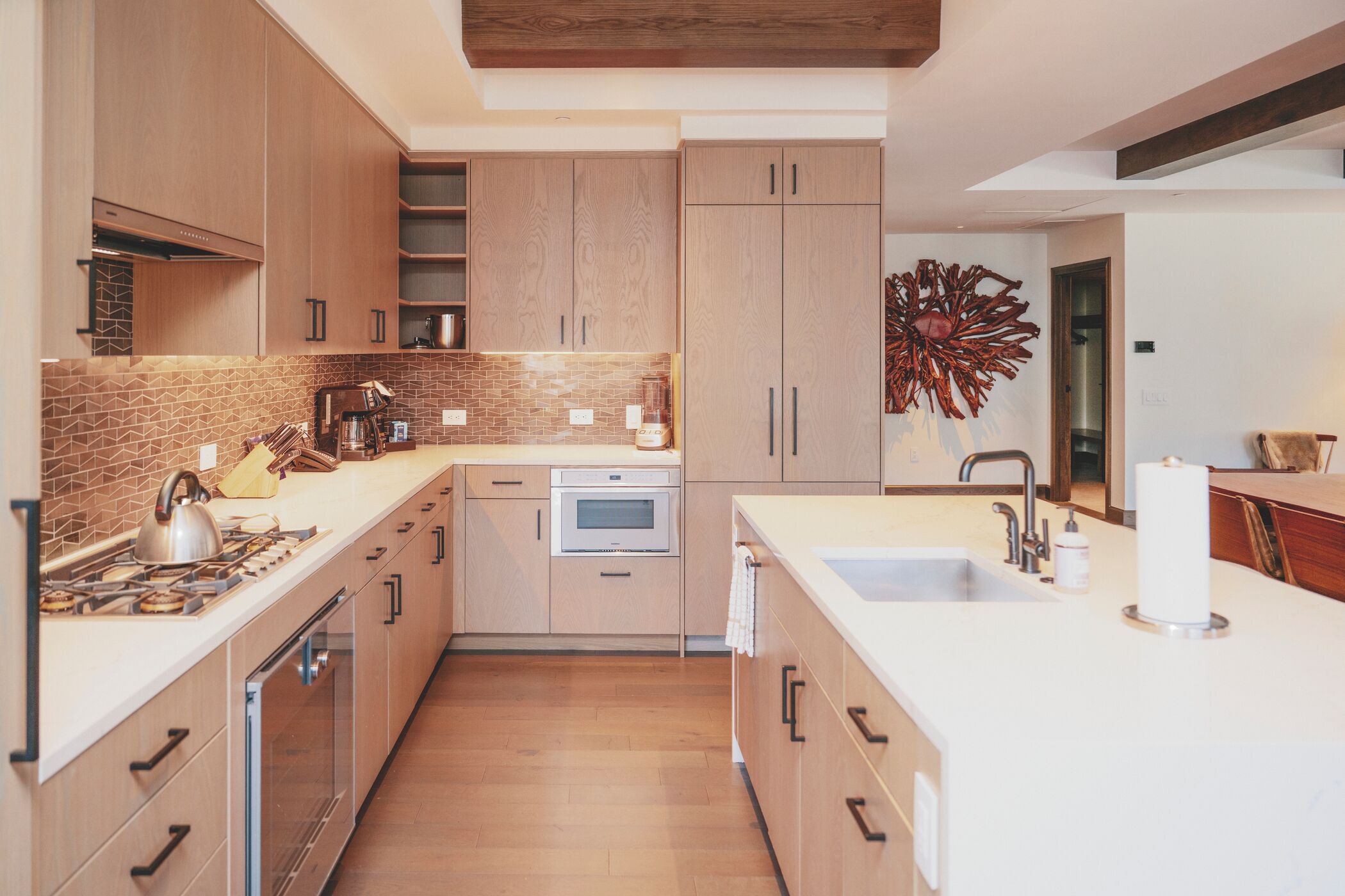 A modern kitchen with wooden cabinets, a tiled backsplash, stainless steel appliances, and a large decorative piece on the wall.