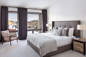Habitación de hotel con una cama grande, una silla, una mesita de noche con lámpara y vistas a un paisaje nevado.