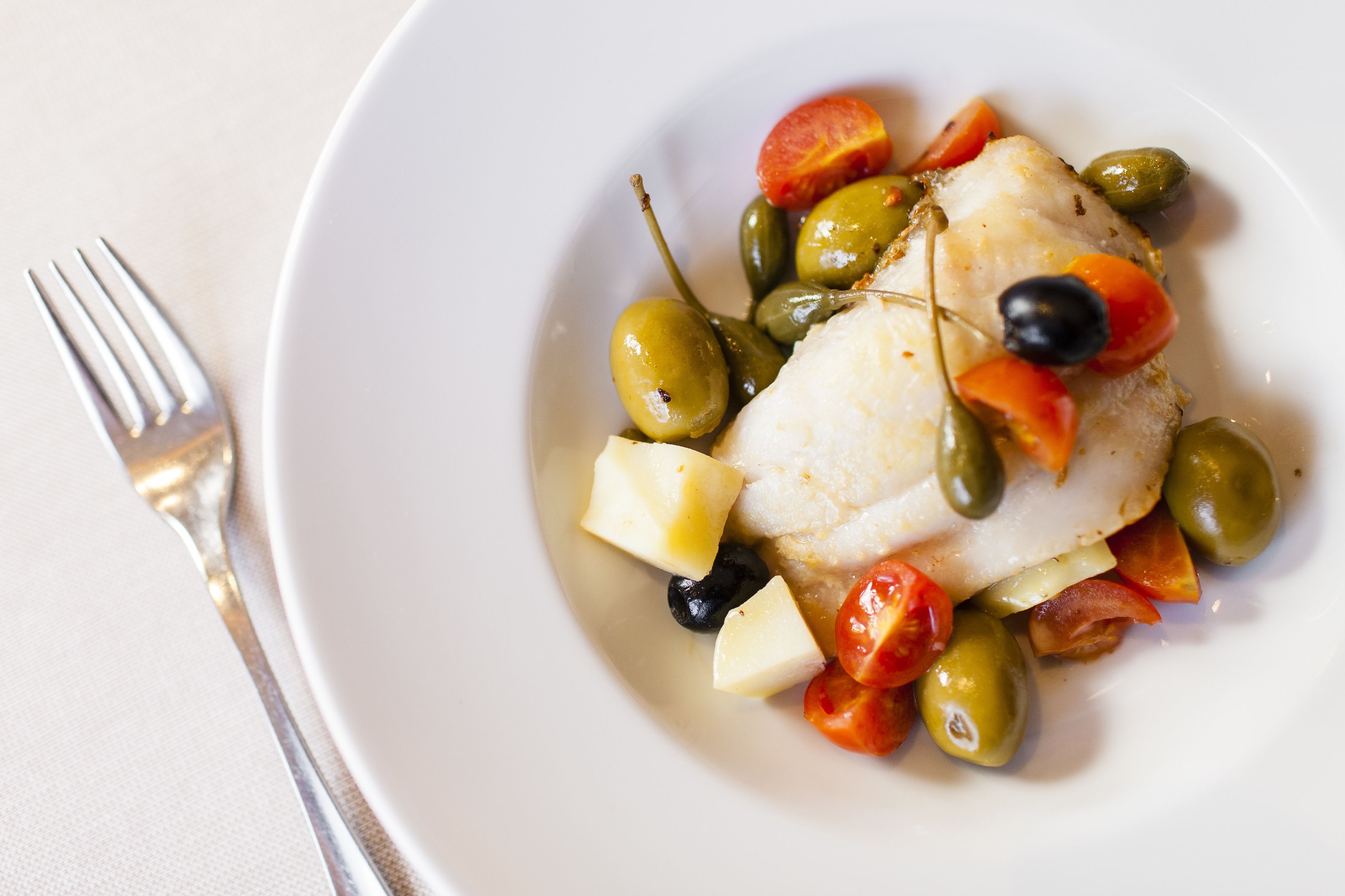 Un plat blanc amb un tros de peix blanc, olives verdes, tomàquets vermells i olives negres.