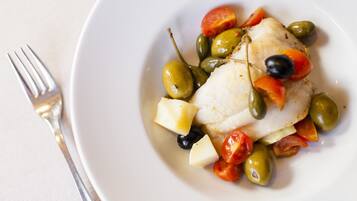 Un plat blanc amb un tros de peix blanc, olives verdes, tomàquets vermells i olives negres.