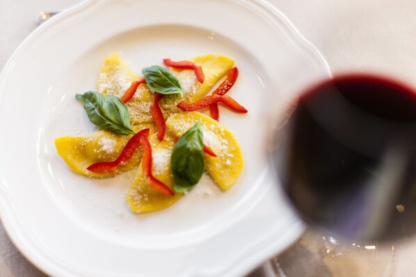 Plato blanco con ravioles adornados con tiras de pimiento rojo y hojas de albahaca, acompañado de una copa de vino tinto.