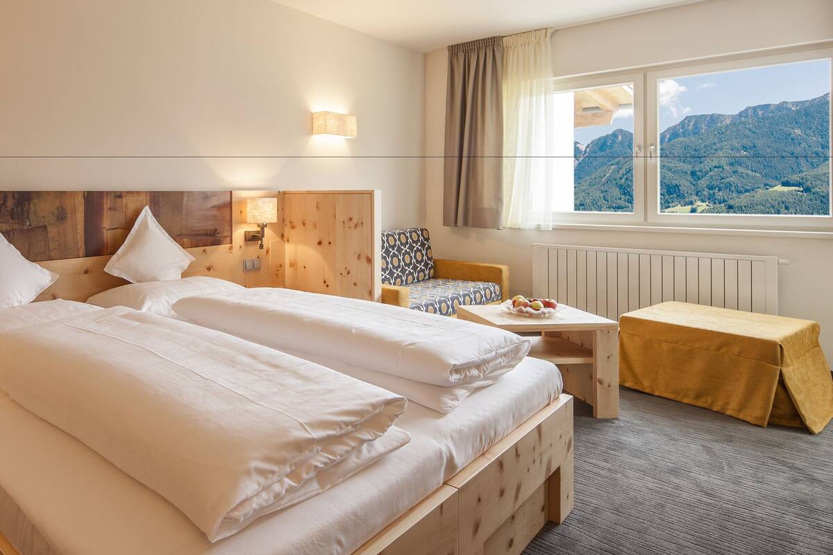 A hotel room with two beds, wooden headboards, a small table, a chair, and a window with a mountain view.