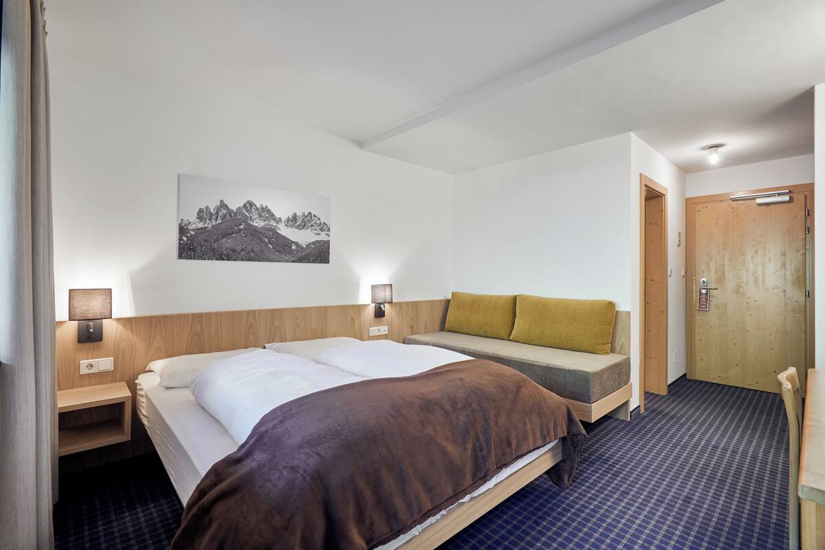 A hotel room with a bed, a sofa, a wall-mounted picture, a wooden door, and a small table with a lamp.