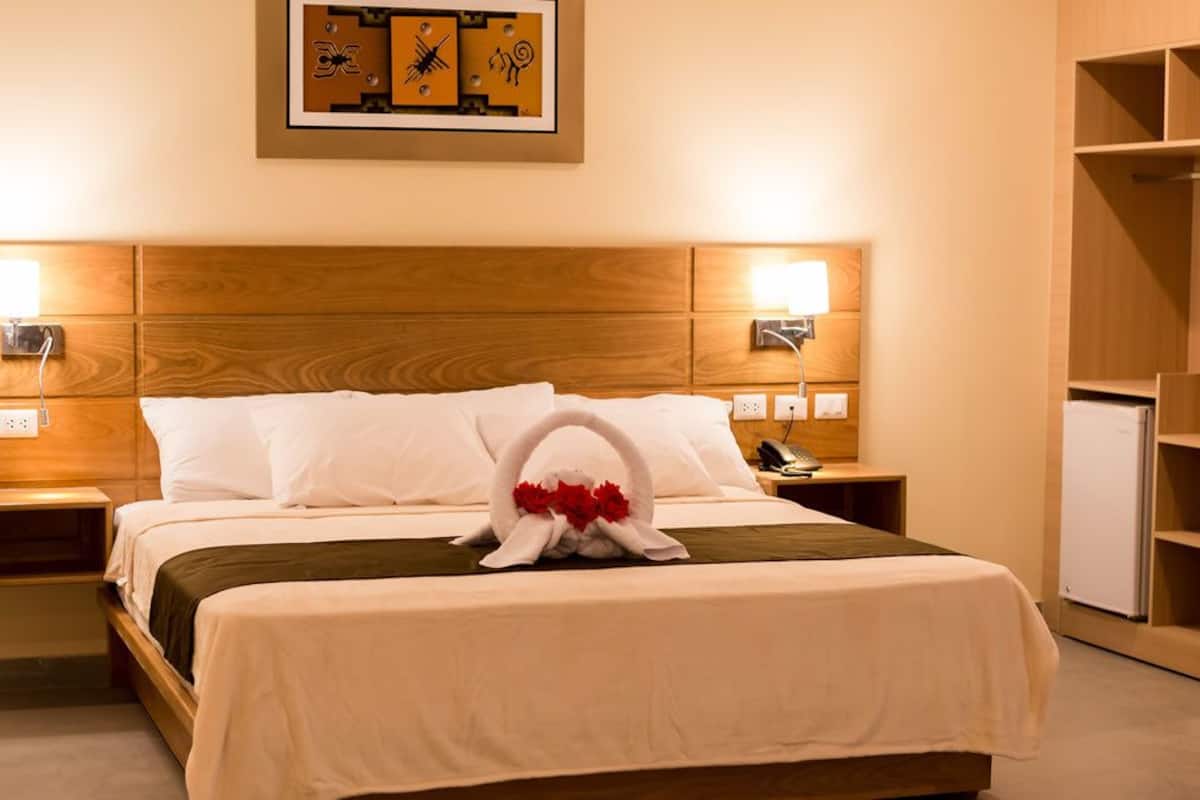 Une chambre d’hôtel avec une tête de lit en bois, deux lampes de chevet, un lit avec du linge de lit blanc, une serviette ornée de fleurs rouges et une petite table avec un téléphone.