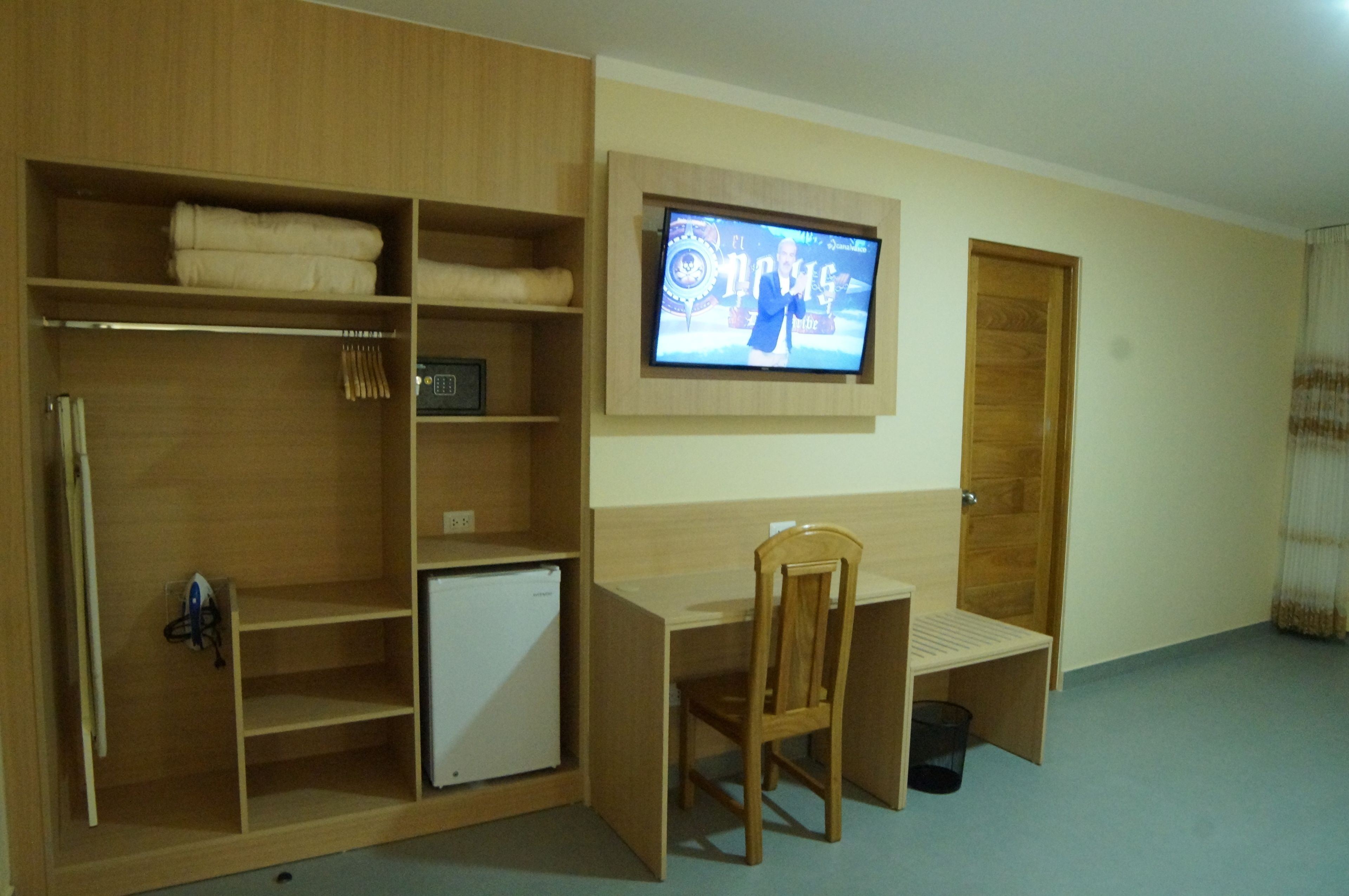 Une pièce comprenant un bureau en bois, une chaise, un téléviseur fixé au mur, une armoire et un petit appareil électroménager blanc.