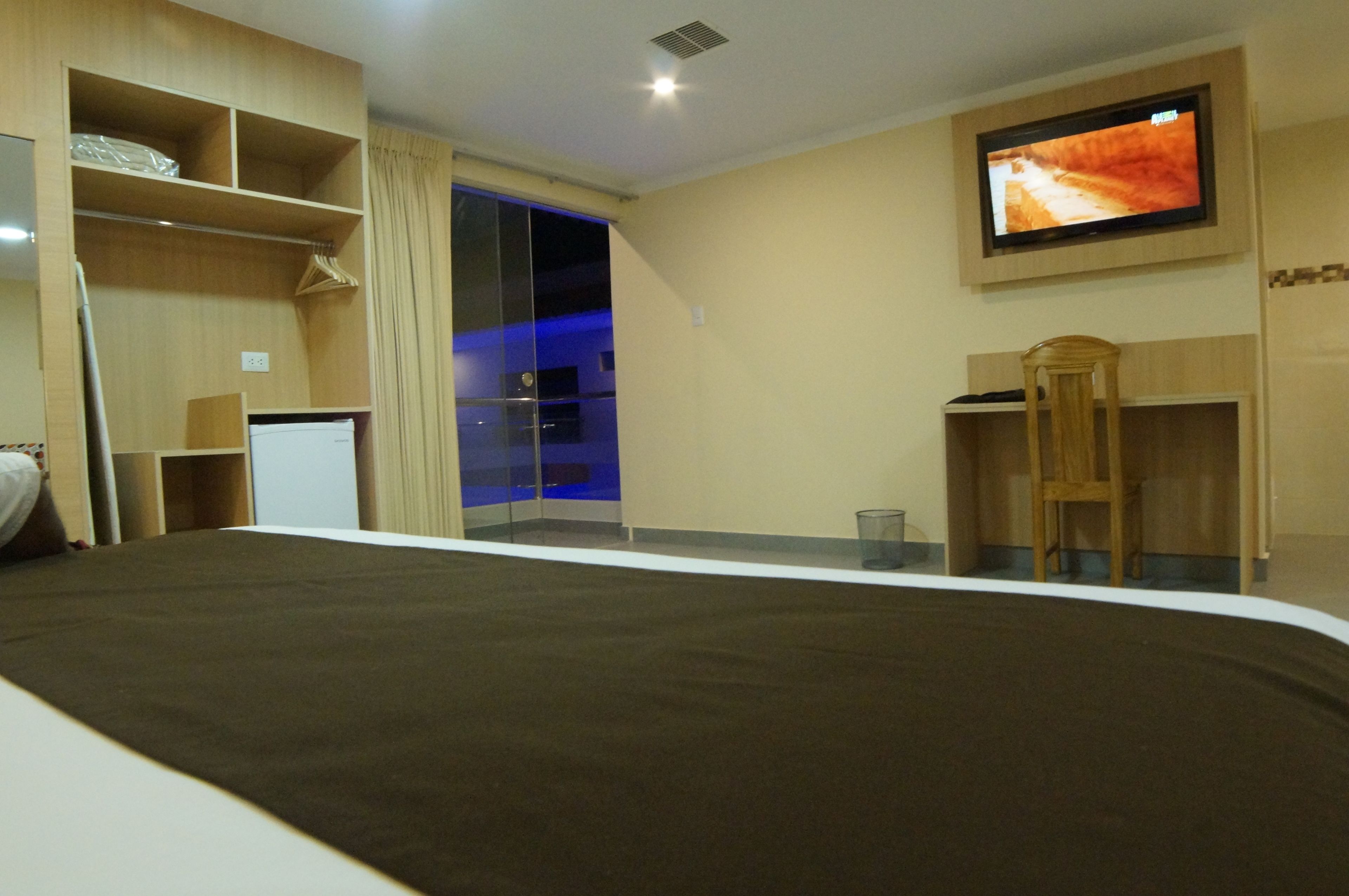 A hotel room with a bed, a TV mounted on the wall, a wooden wardrobe, and a glass door leading to a balcony.