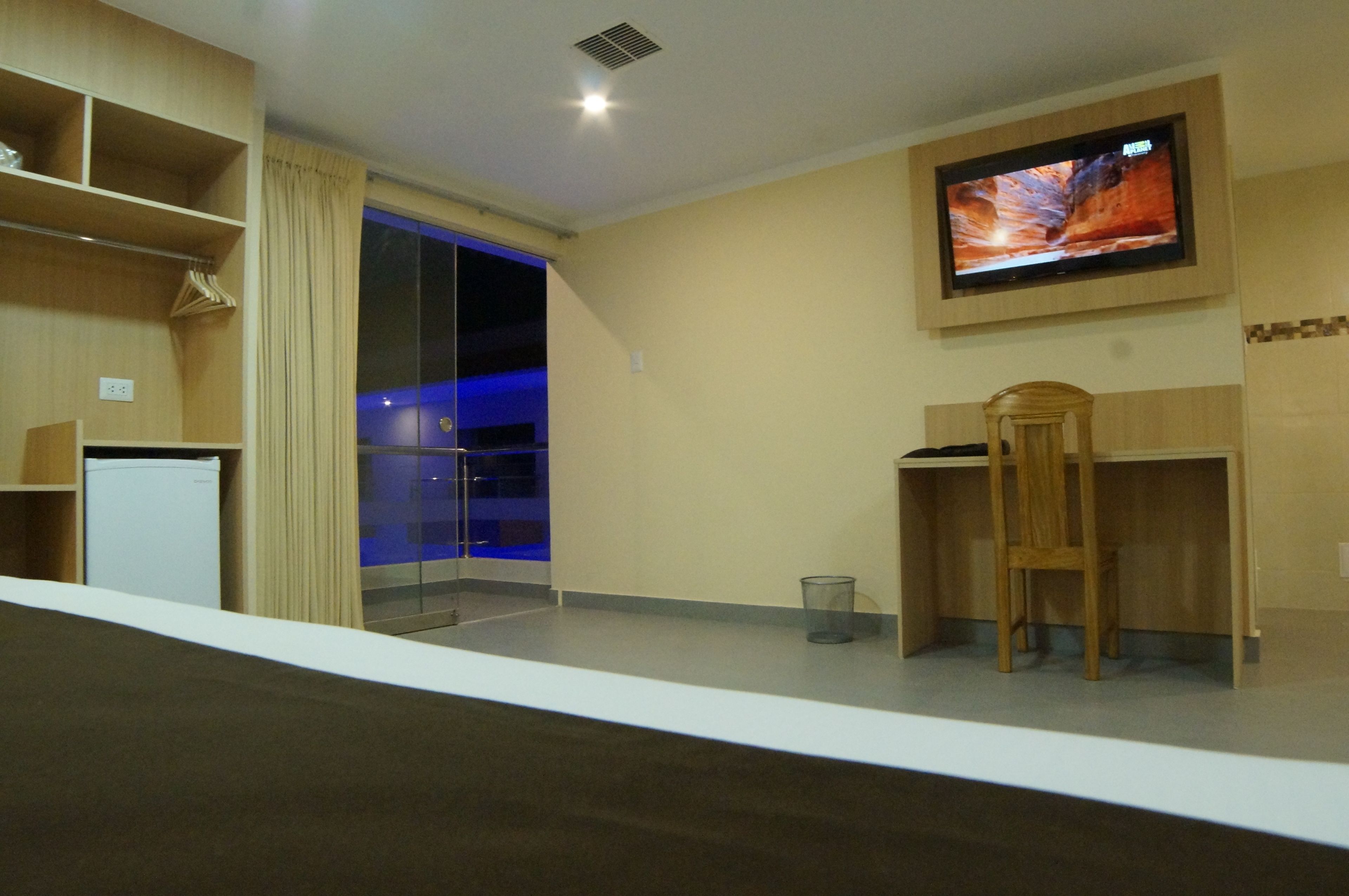 Habitación de hotel con televisión montada en la pared, un escritorio pequeño, una silla y una puerta de vidrio que da a un balcón.