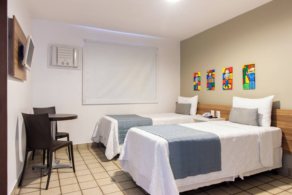 Habitación de hotel con dos camas, una mesita con una silla, televisor montado en la pared y cuadros de colores en la pared.