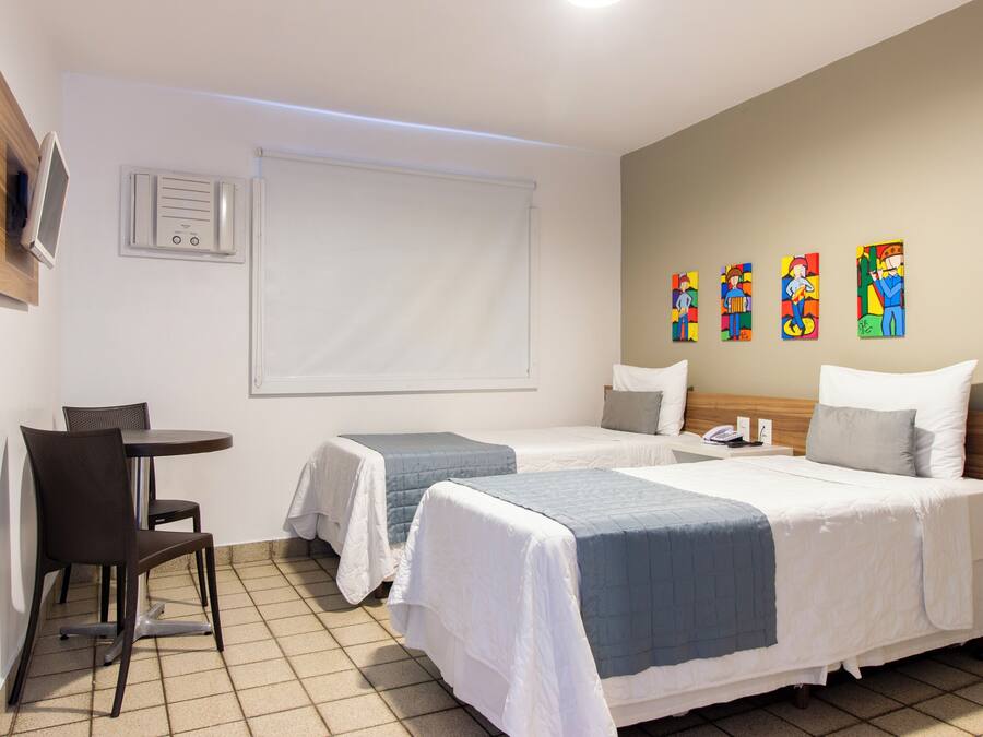 A hotel room with two beds, a small table with a chair, a TV mounted on the wall, and colorful artwork on the wall.