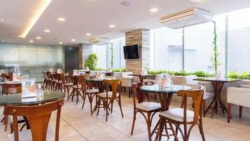 Un restaurante moderno con mesas y sillas redondas, un televisor de pantalla plana en la pared y ventanas amplias con plantas.