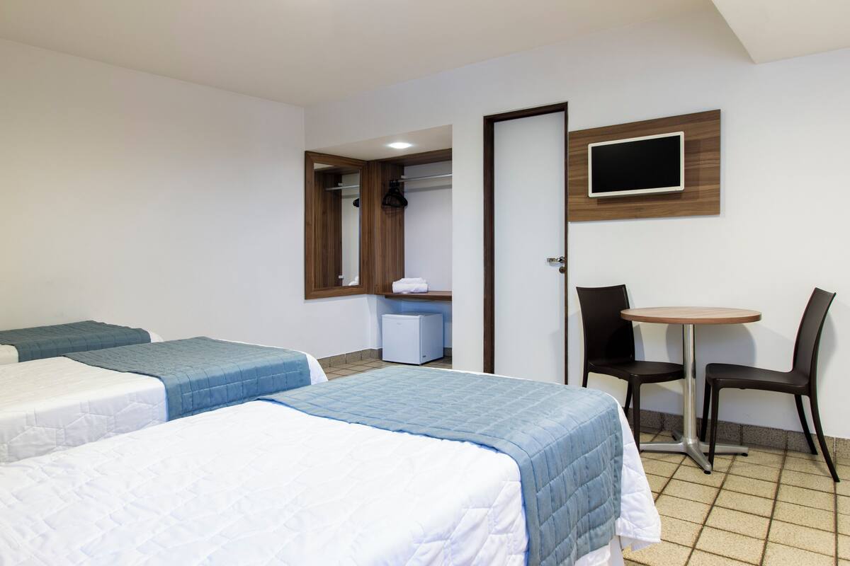 Habitación de hotel con dos camas, una pequeña mesa con sillas, un televisor montado en la pared y un baño visible a través de una puerta abierta.