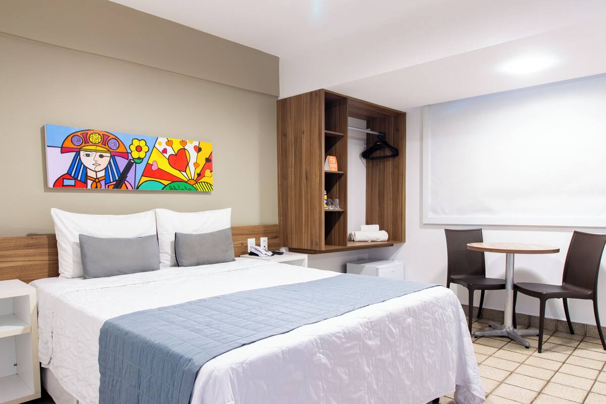 Habitación de hotel con una cama grande, un armario de madera, una mesita con dos sillas y un cuadro en la pared.