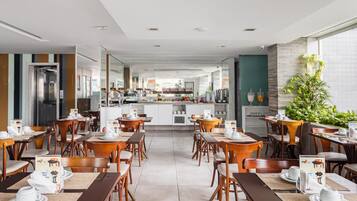 Un comedor de hotel moderno con mesas y sillas de madera, una cocina central y vistas a la ciudad.