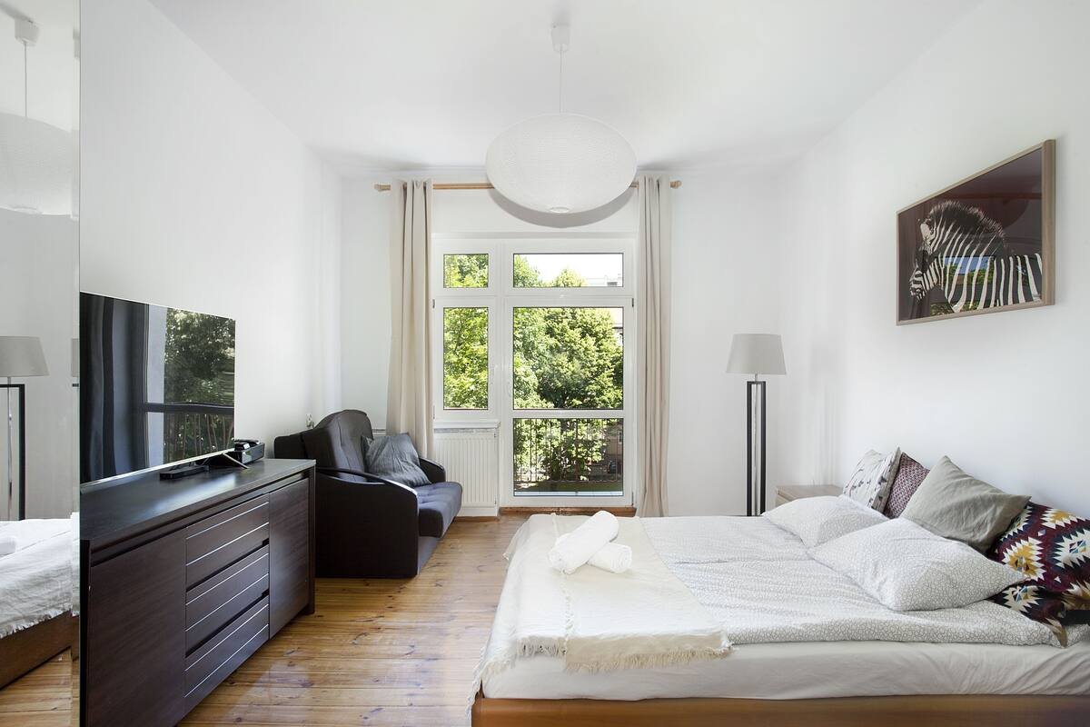 A modern bedroom with a large bed, a dresser, a TV, and a window with a view of trees.