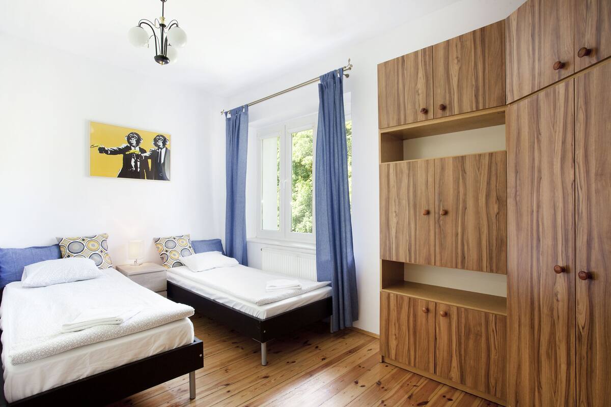 A bedroom with two beds, a wooden wardrobe, a window with curtains, and a painting on the wall.