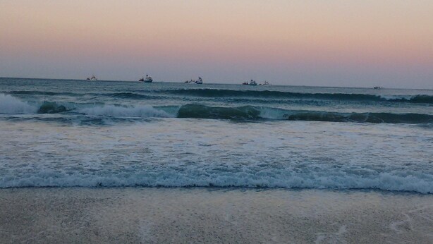 Een strand met golven en schepen in de verte.