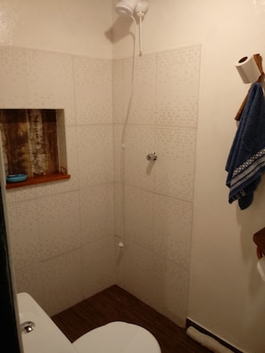 A small, tiled shower area with a white showerhead, a wooden shelf, and a blue towel hanging on the wall. - Casinha Charmosa (Florianópolis)