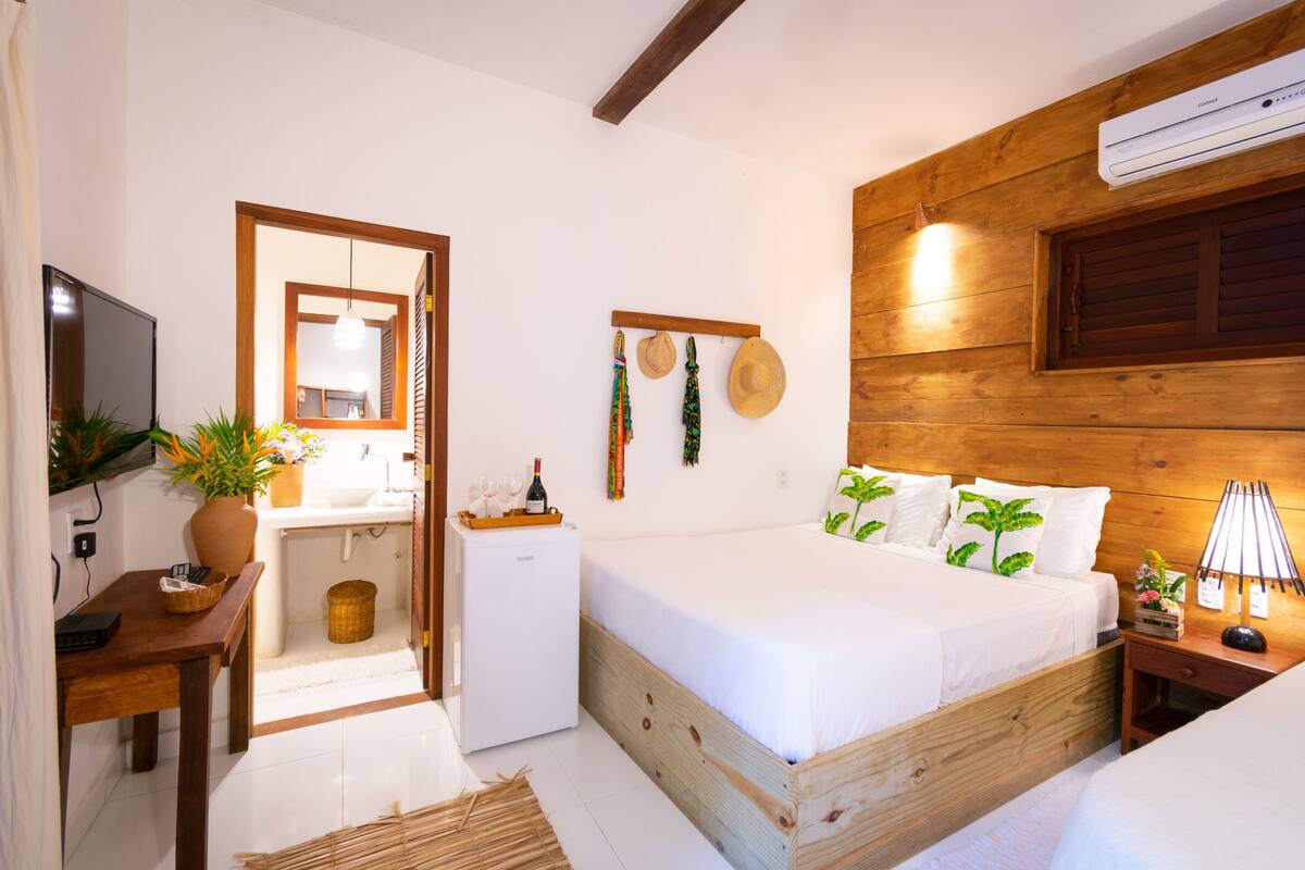 A cozy bedroom with a wooden headboard, a bed with white bedding, a bedside table with a lamp, and a small desk with a computer and a plant.