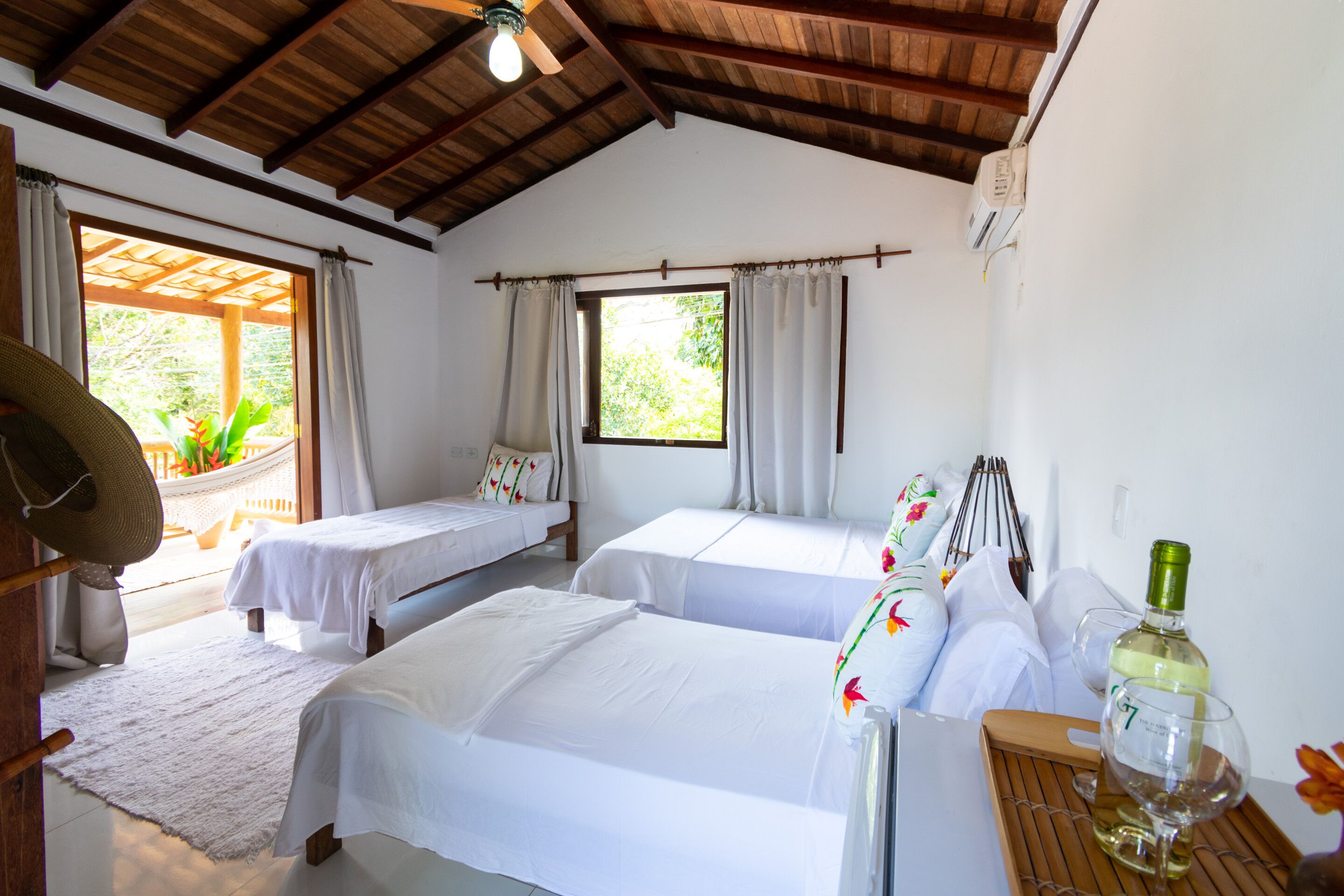 A room with two beds, a wooden ceiling, a window with curtains, a hat on a chair, and a bottle of wine on a tray.