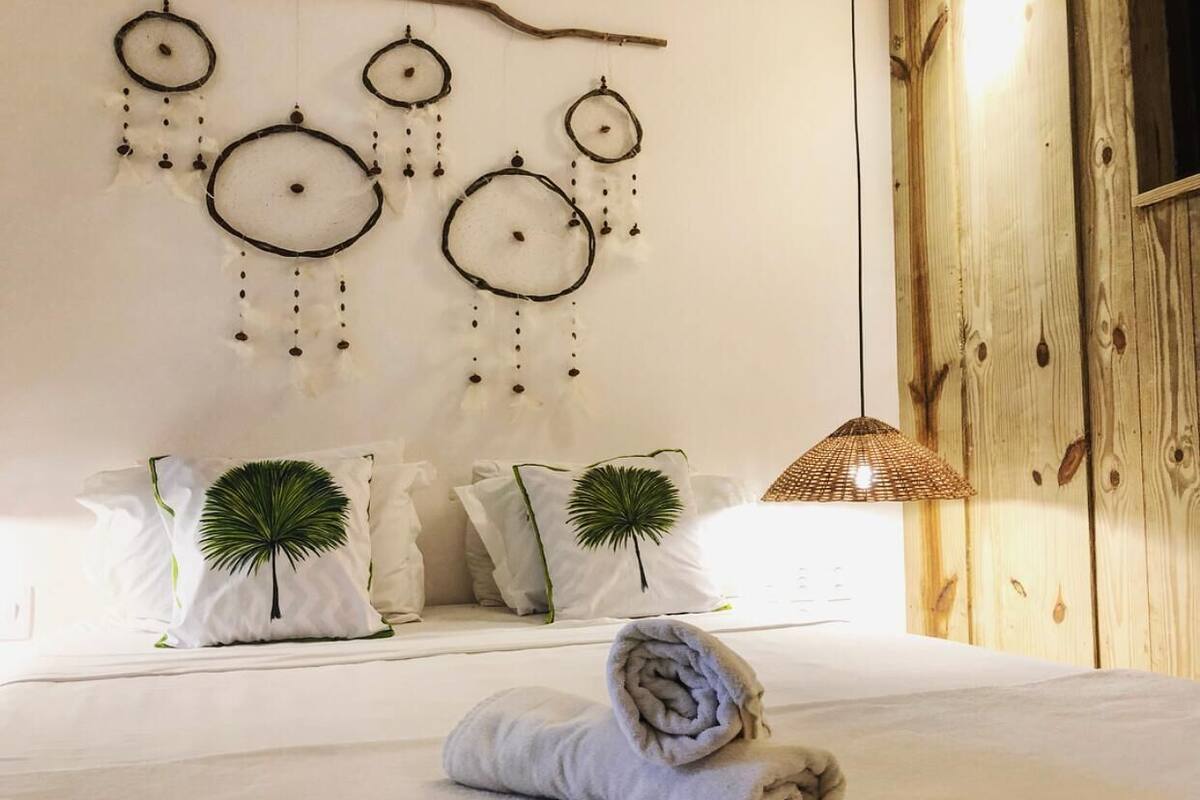 A neatly made bed with a rolled towel, a wall-mounted dreamcatcher, and two decorative pillows with palm leaf designs.