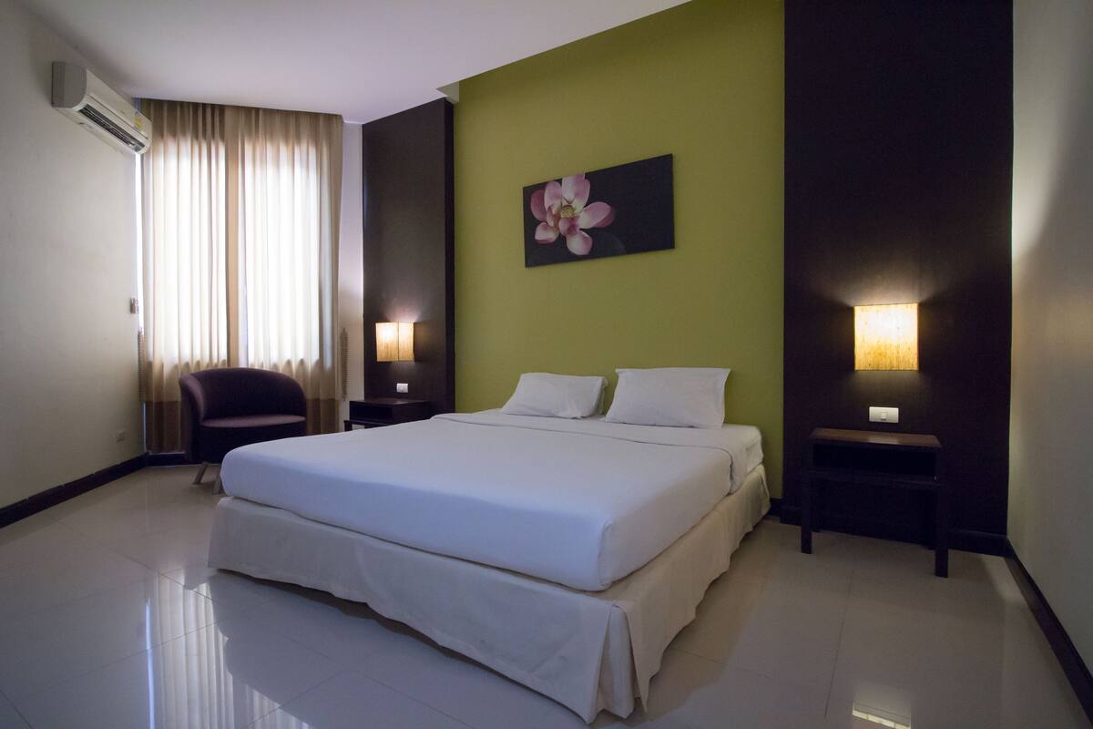Habitación de hotel con una cama grande, dos mesitas de noche con lámparas, una silla, aire acondicionado en la pared y un cuadro de flores.