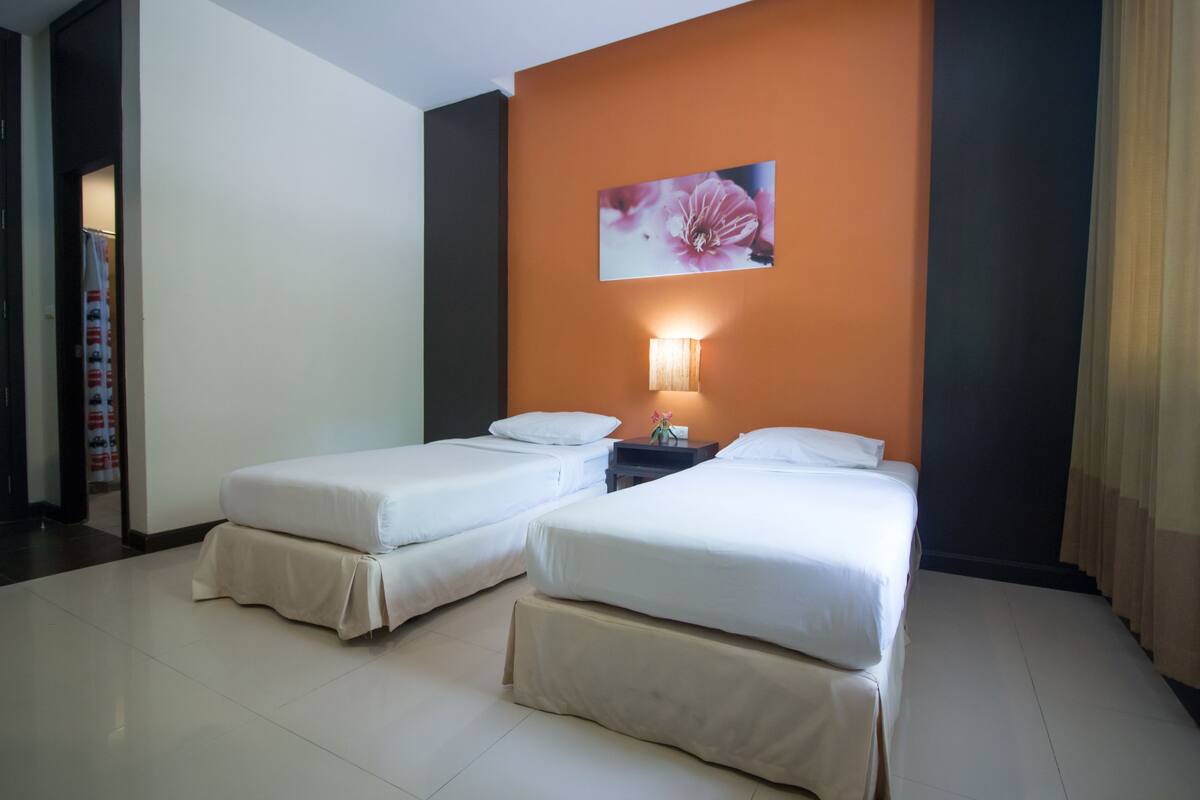 Habitación de hotel con dos camas individuales, una mesita de noche con lámpara y un adorno floral en la pared.