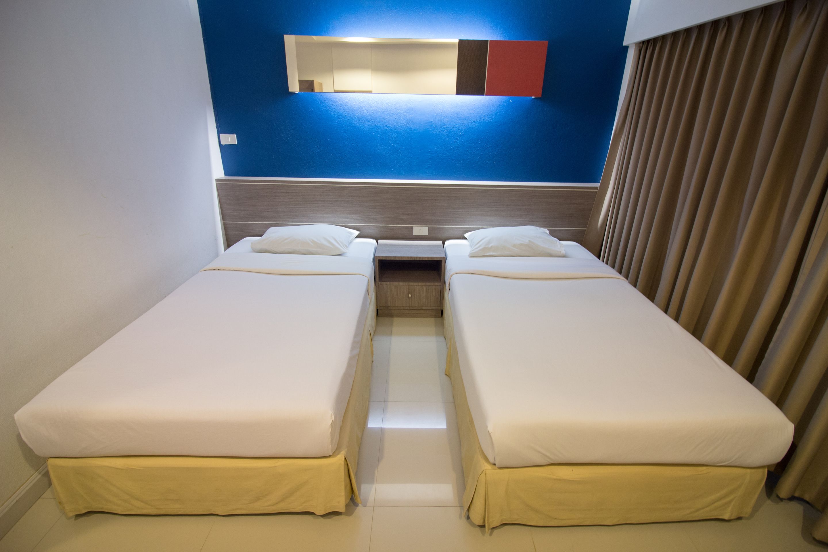 Quarto de hotel com duas camas de solteiro, uma mesinha, um espelho e uma janela com cortinas.