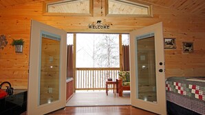 A wooden cabin interior with a wooden deck, a welcome sign, a bed with a quilt, and a small table with chairs.