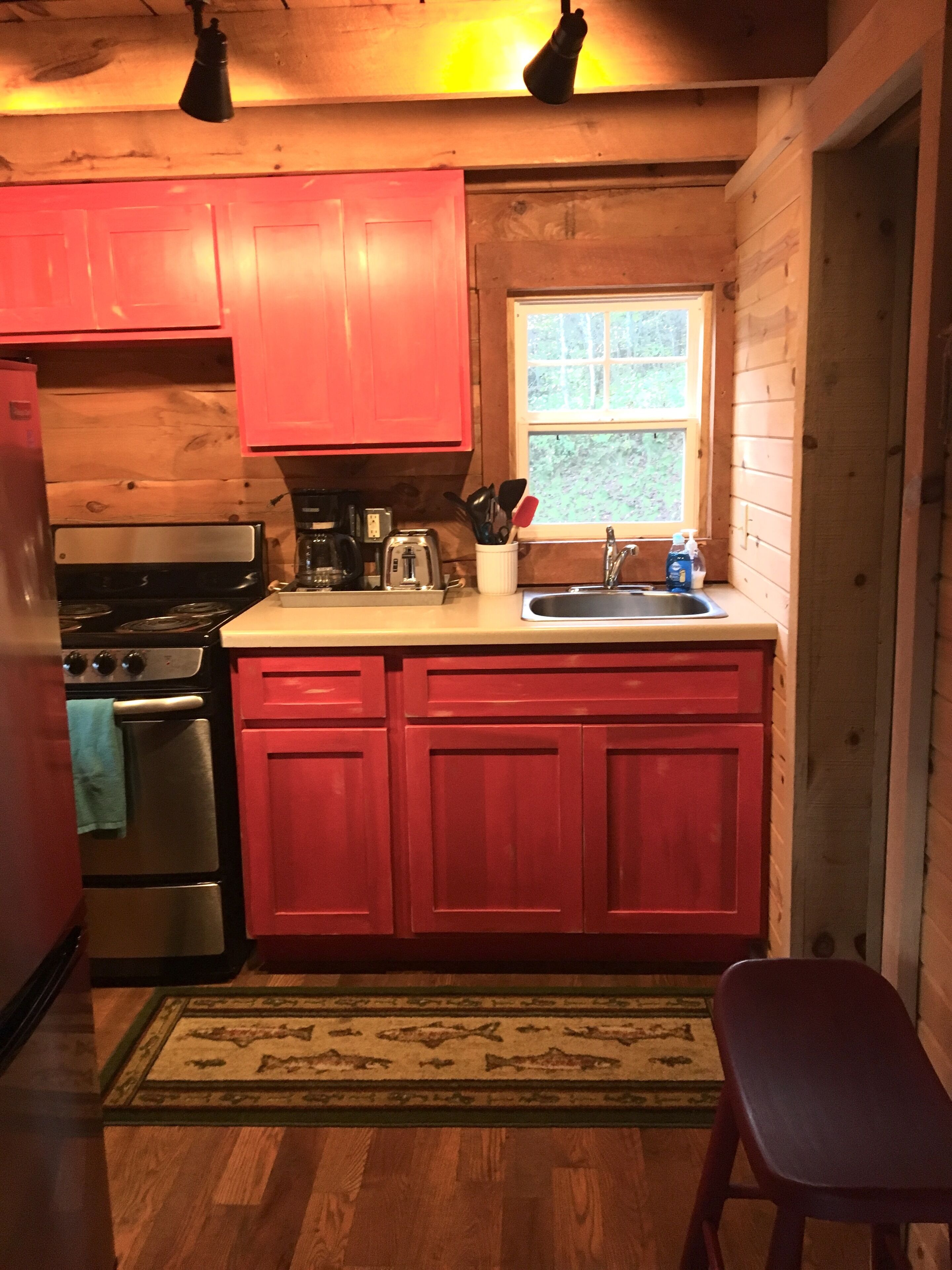 Cocina compacta con paredes de madera, estufa de acero inoxidable, armarios rojos y una ventana.