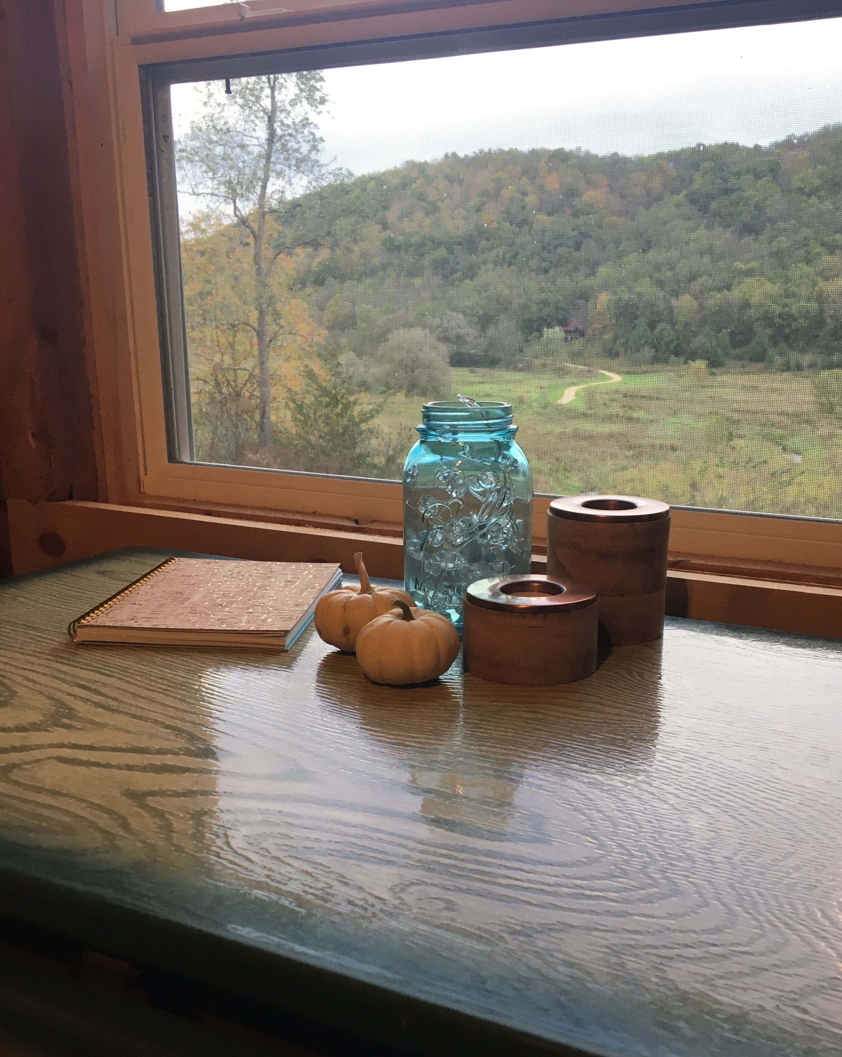 Un tavolo di legno con un quaderno, due piccole zucche, un barattolo di vetro blu e due candelabri in rame vicino a una finestra con vista su un paesaggio verde.
