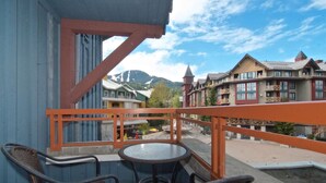 A balcony with wicker chairs and a small round table, overlooking a town with colorful buildings and a mountain in the background.