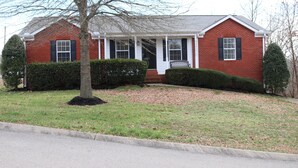 A two-story brick house with a front porch, a small front yard, and a tree in front.
