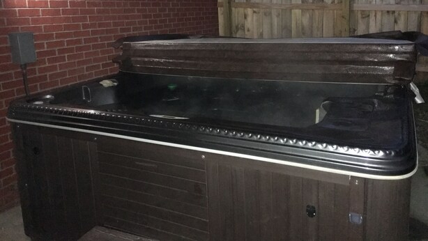 A hot tub with a wooden exterior, positioned against a brick wall and a wooden fence.
