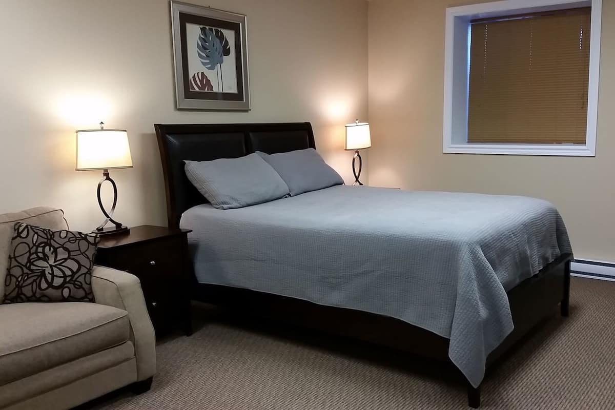 A bedroom with a bed, two bedside lamps, a framed artwork, a window with blinds, and a chair.