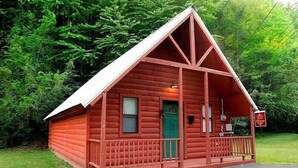 A small, red log cabin with a green door, a porch, and a sign indicating it is a guesthouse. - Gatlinburg Cabin Free Tickets, Heart Whirlpool, Hot Tub, King Bed, Fast Internet (Gatlinburg)
