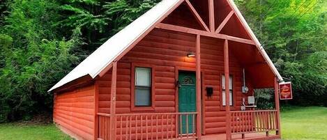 A small, red log cabin with a green door, a porch, and a sign indicating it is a guesthouse.
