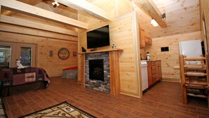 A cozy log cabin interior with a stone fireplace, wooden walls, a television mounted above it, and a dining area with wooden cabinets. - Gatlinburg Cabin Free Tickets, Heart Whirlpool, Hot Tub, King Bed, Fast Internet (Gatlinburg)