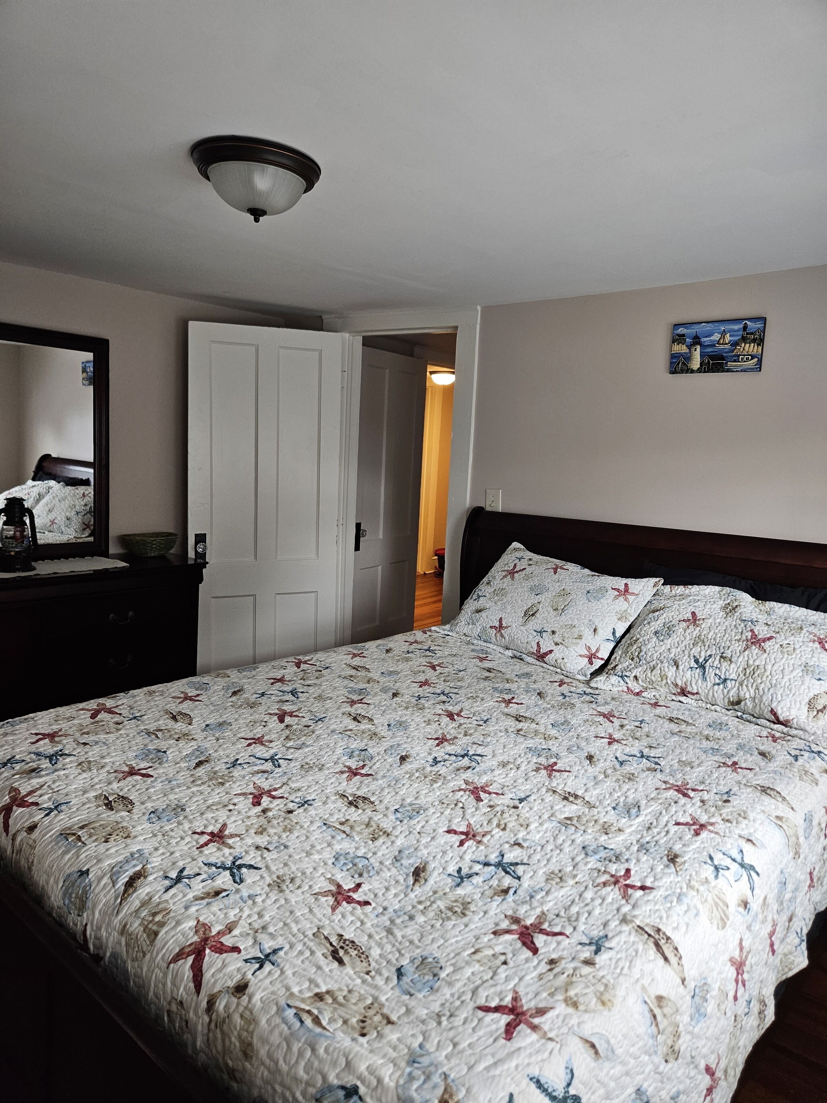A bedroom with a bed, a dresser, a mirror, a door, and a wall with a painting.