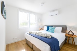A bedroom with a bed, a window with blinds, a bedside table with a lamp, and an air conditioning unit on the wall. - Hampton Lookout Rooftop Terrace with Bay Views (Hampton)
