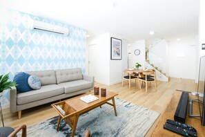 A modern living room with a grey sofa, a coffee table, and a dining area with a wooden table and chairs. - Hampton Lookout Rooftop Terrace with Bay Views (Hampton)