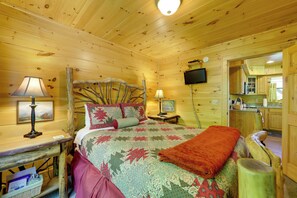 A wooden cabin bedroom with a bed, a desk with a lamp, a television, and a view of the kitchen. - Hot Tub, Fire Pit & Mtn Views: Bryson City Escape (Bryson City)