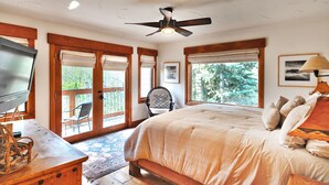 A bedroom with a large bed, a ceiling fan, a TV, and a chair by the window.