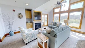 A spacious living room with a fireplace, a large window with a view, and a ceiling fan. - Escape to OBX!! at Hamilton Cay 2 Bedroom Premium Condo (Kill Devil Hills)
