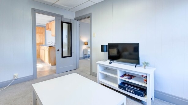Un salon moderne avec un téléviseur à écran plat posé sur un meuble blanc, une table basse blanche et une porte-fenêtre donnant sur un espace cuisine.