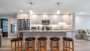 Une cuisine moderne dotée d’un îlot central, de tabourets de bar, d’appareils électroménagers en acier inoxydable et de luminaires suspendus.
