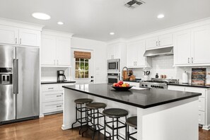 A modern kitchen with white cabinets, a stainless steel refrigerator, a large island with stools, and a gas stove. - Bevin's Haus | Beautiful Interior | Terrific Backyard (Fredericksburg)