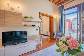 Una sala de estar moderna con televisor, armarios de madera, una mesa de centro de vidrio y un balcón con vistas a edificios.