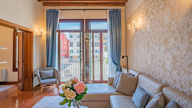 A room with a wooden ceiling, a sofa, a chair, a table with flowers, and a view of buildings through a glass door. - Ca' Del Monastero 1 Collection with Balcony (Venice)