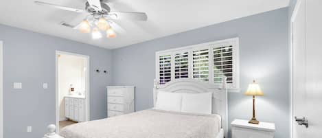 Une chambre à coucher comprenant un lit, une table de chevet, une commode, un ventilateur de plafond et une fenêtre avec des stores.
