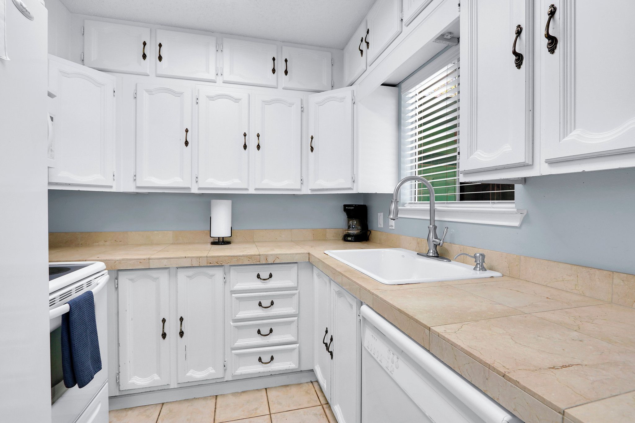 A kitchen with white cabinets, a sink, a coffee maker, and a window with blinds.